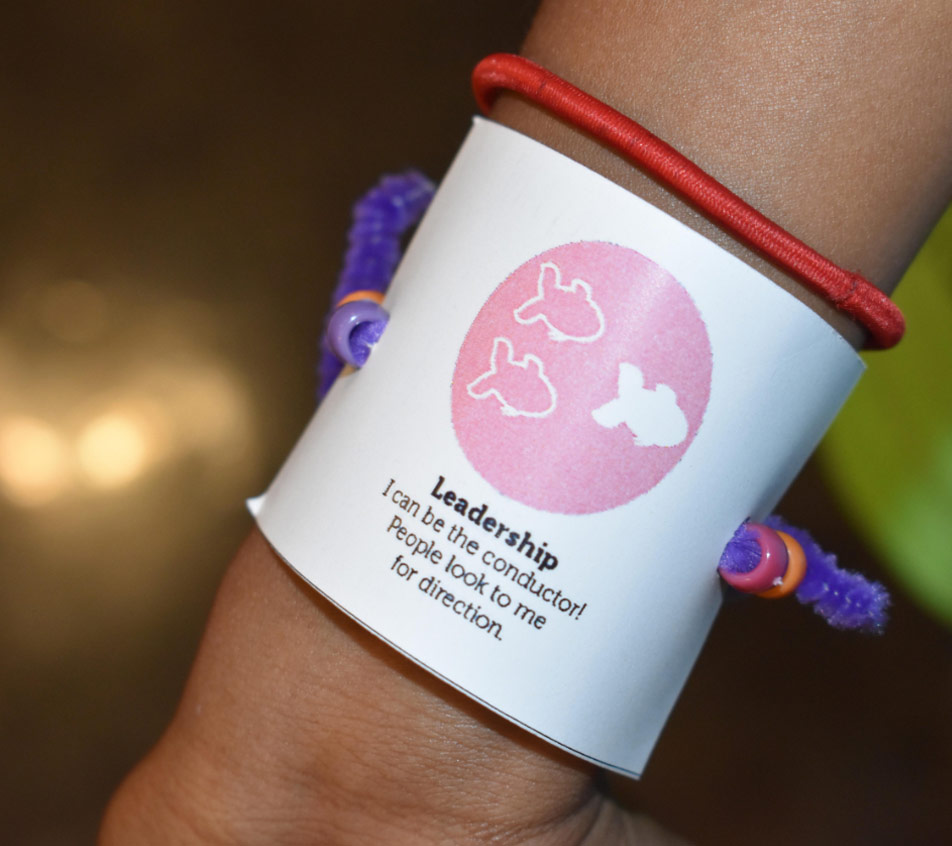 Zoomed in photo of a child's wrist with a red hair tie and a purple bracelet with a white piece of paper with the Leadership character strength and logo on it